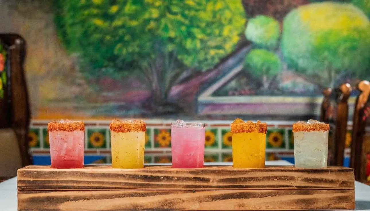 Five salt-rimmed margaritas on a wooden board in front of a painted garden mural