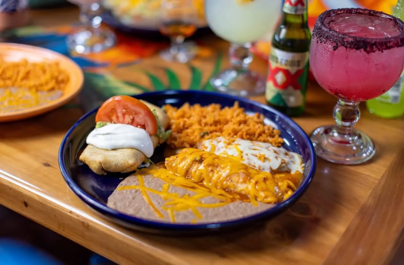 A lunch plate of enchiladas with rice and refried beans on a blue plate, a pink margarita and a Dos Equis behind