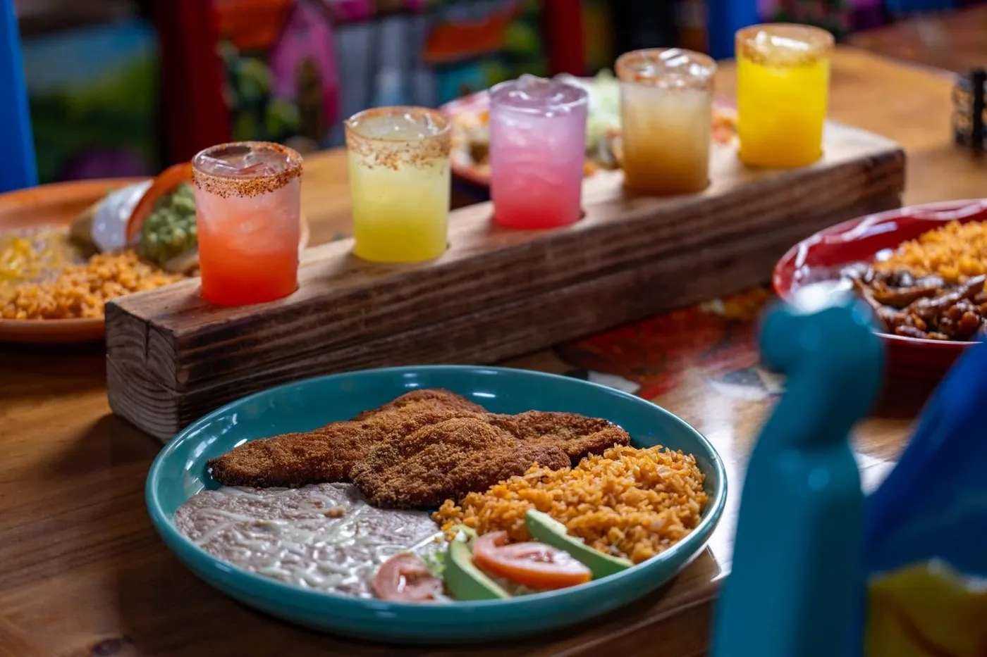 A flight of five salt-rimmed margaritas on a wooden board behind a breaded fish plate with rice and beans