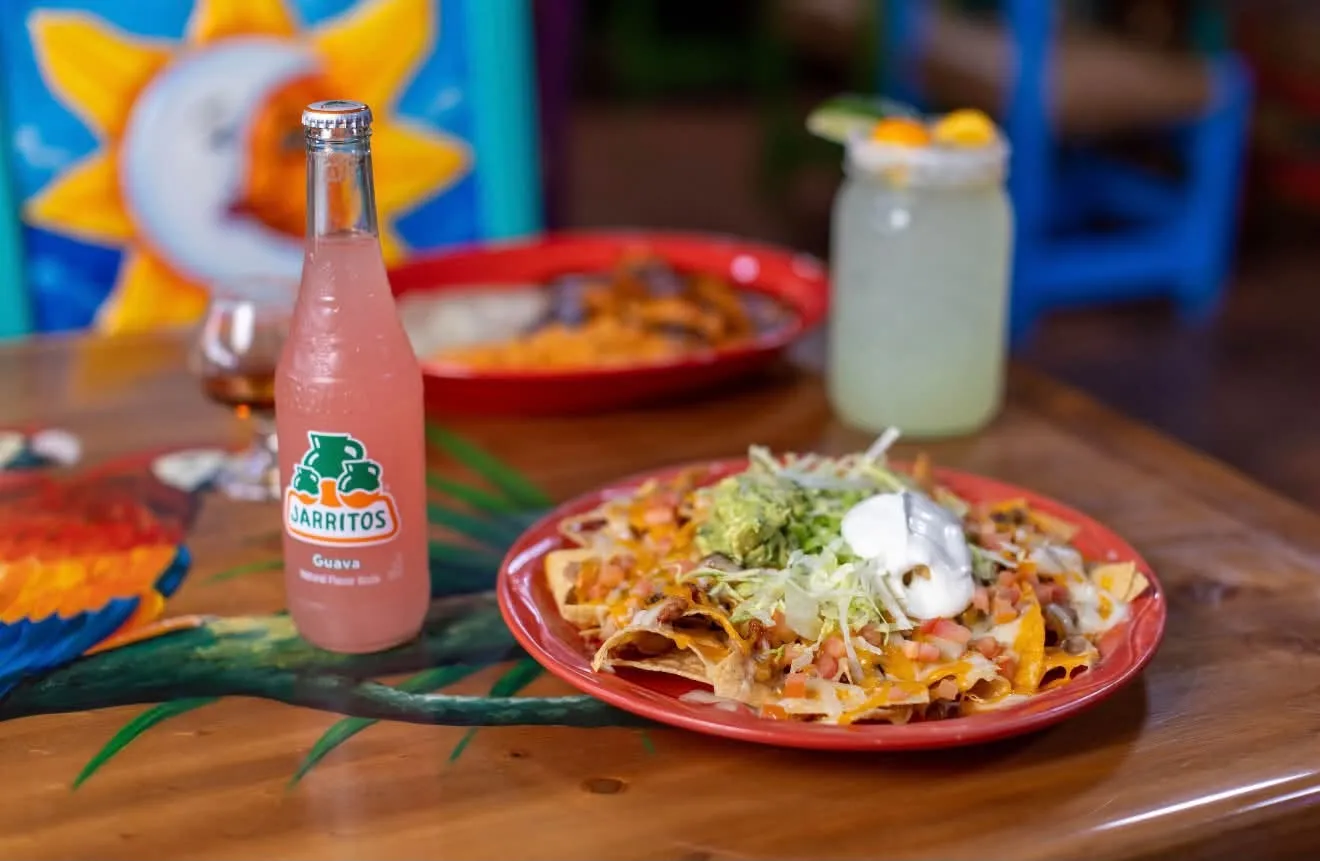 Loaded nachos with guacamole and sour cream on a red plate, a guava Jarritos beside them