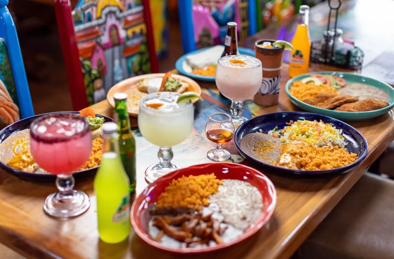 A table full of plates, margaritas and a Jarritos in front of La Roca’s painted chairs